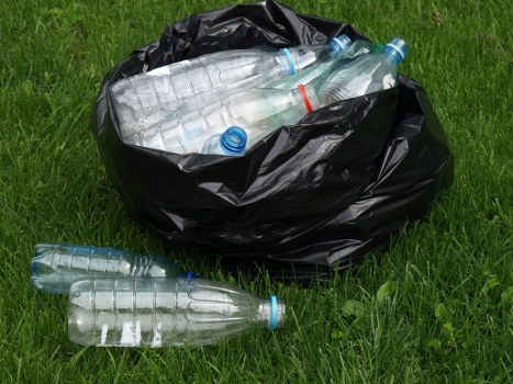 A black plastic rubbish bag placed on a grassy outdoor area, filled with several clear plastic bottles with blue caps and some red markings, most of which are partially visible at the top of the bag and some lying on the grass nearby. The bag appears to be full and loosely closed, with a few bottles spilling out onto the ground. The scene suggests a waste removal activity typically handled by professional rubbish clearance services such as Office Clearance Belmont, located in the Belmont or neighbouring town area. The grassy environment is lush and green, providing a natural contrast to the clear plastic bottles and the dark rubbish bag. The bottles vary in size and shape, indicating they are likely discarded beverage containers, and the overall scene is clean, orderly, and indicative of waste collection or disposal efforts in a residential or semi-urban setting, which is common for rubbish removal providers operating in the Belmont postcode area.