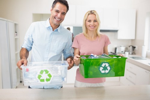 A man and a woman standing in a modern kitchen, each holding a transparent plastic recycling bin with a green recycling symbol printed on the side; the man’s bin appears to contain clear plastic bottles, while the woman’s bin is solid green and empty; both individuals are smiling; the kitchen background features white cabinetry, a stainless steel kettle, and various small appliances, with natural lighting illuminating the space; this image relates to rubbish and recycling services offered by Office Clearance Belmont in Belmont, possibly within the postcode area related to the town, highlighting the importance of proper waste separation and disposal.