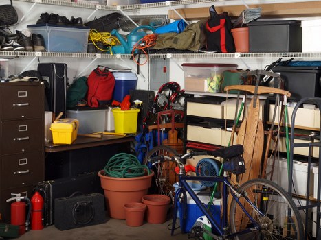 The image shows a cluttered garage or storage area filled with various household and outdoor items. In the foreground, there are several empty terracotta plant pots of different sizes stacked on the floor. A blue bicycle with a black saddle and a small black bag attached to the frame leans against a shelving unit. Surrounding the bike, there are hoses, a red and black portable fan, and a black plastic storage box. To the left, a black metal filing cabinet and a yellow plastic container sit on a work surface, with a red tool box and a gray toolbox nearby. Above, metal shelving units hold plastic storage bins, cleaning supplies, and miscellaneous tools, with some boxes and bags visibly placed on the shelves. The scene is densely packed, implying a typical household or garage clutter, reflecting the type of rubbish that a rubbish removal service like Office Clearance Belmont might handle during a clearance in the Belmont area. The lighting appears to be natural or from overhead fixtures, highlighting the variety of objects and the disorganized state of the storage space.