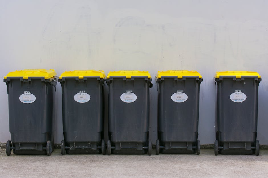 A row of five large, dark grey wheelie bins with bright yellow lids lined up along a smooth, light grey concrete surface against a plain, off-white wall. Each bin features a white oval label with blac