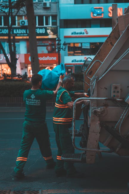 A worker dressed in a high-visibility orange vest, black jacket, and reflective trousers is standing on a pavement next to a blue truck, holding an orange traffic cone in front of a large gray waste s