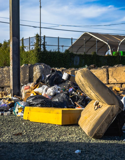 A person crouching on a paved outdoor surface, wearing light blue jeans and white sneakers, holding a small collection of discarded rubbish in their right hand. The rubbish includes a white plastic or