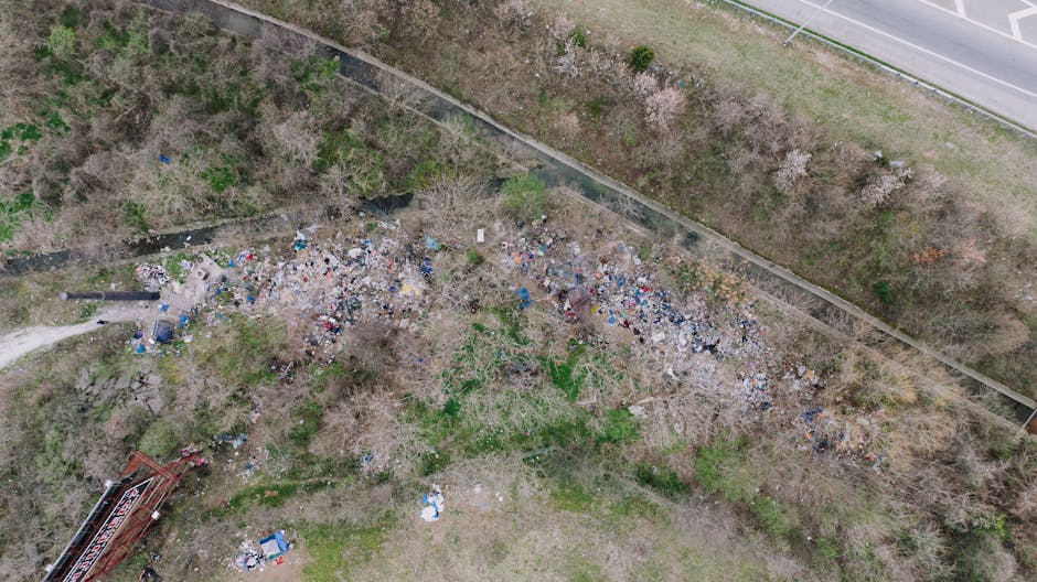 A large pile of mixed household waste and rubbish, including crumpled cardboard boxes, plastic bags, paper, and discarded packaging, overflowing from a metal skip with a blue lid in an urban parking a